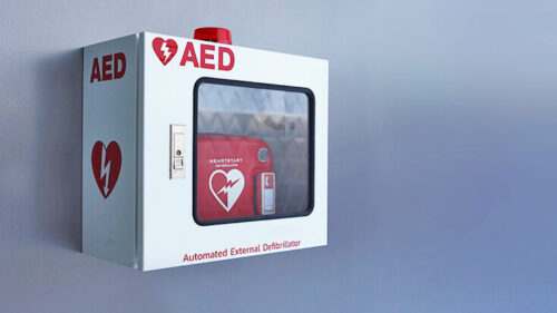 A white wall-mounted box labeled AED with a red heart and lightning bolt symbol contains an automated external defibrillator and a red kit inside, visible through a clear window.